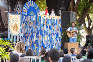 Músico segurando violão em um palco montado ao ar livre durante apresentação artística com show de fantoches na FLIB